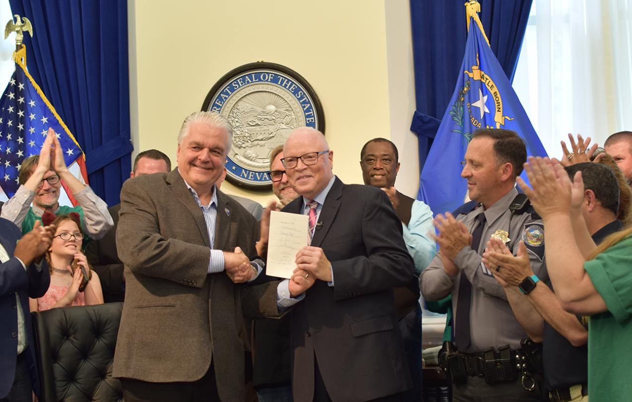 A Crowd of people applaud while two men in the center shake hands, while one displays a signed document